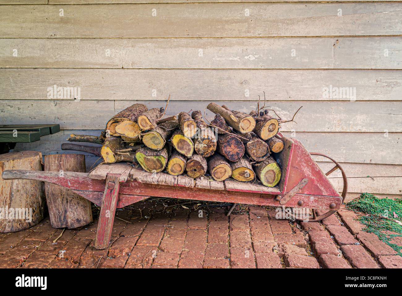 Una scena rustica che mostra una carriola in legno d'epoca piena di tronchi tritati, posta contro un muro di legno intempestivo. L'ambientazione evoca un senso di si Foto Stock