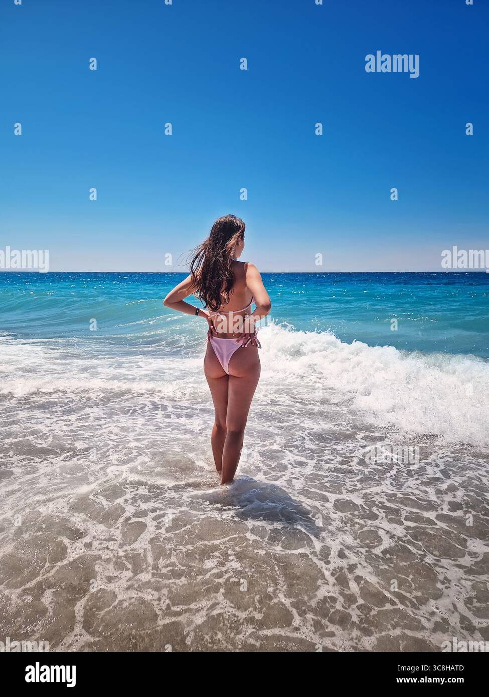 Vista posteriore la bella donna in bikini si erge tra le onde schiumose del mare azzurro, di fronte all'ampio orizzonte sotto un cielo estivo senza nuvole. Estate Foto Stock