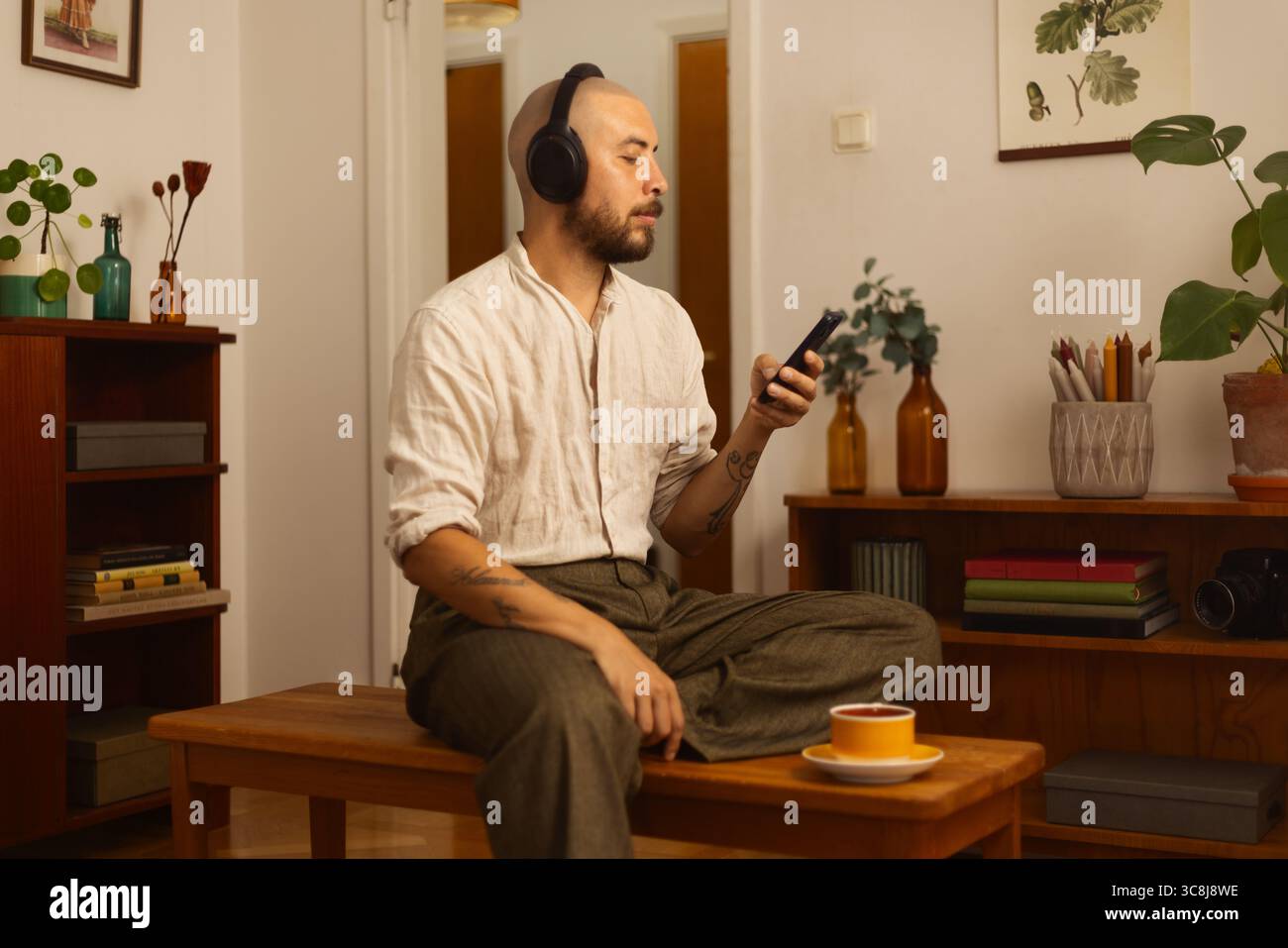 Uomo seduto a gambe incrociate su una panchina, con cuffie e smartphone in una stanza accogliente ed elegante. Un momento di calma di concentrazione digitale e relax. Foto Stock