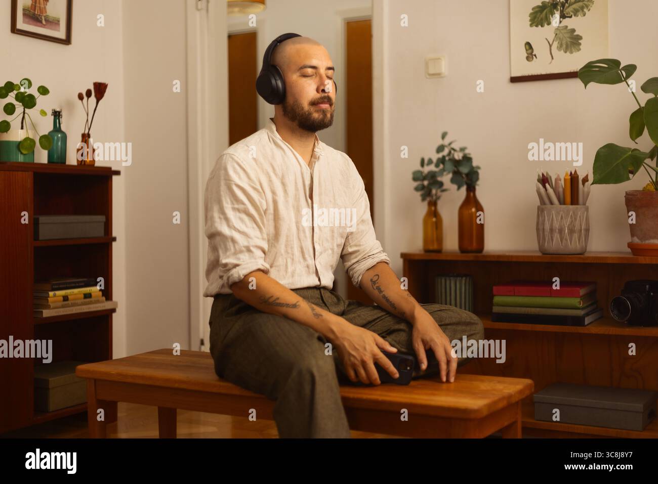 Uomo seduto a gambe incrociate su una panchina, con cuffie e smartphone in una stanza accogliente ed elegante. Un momento di calma di concentrazione digitale e relax. Foto Stock