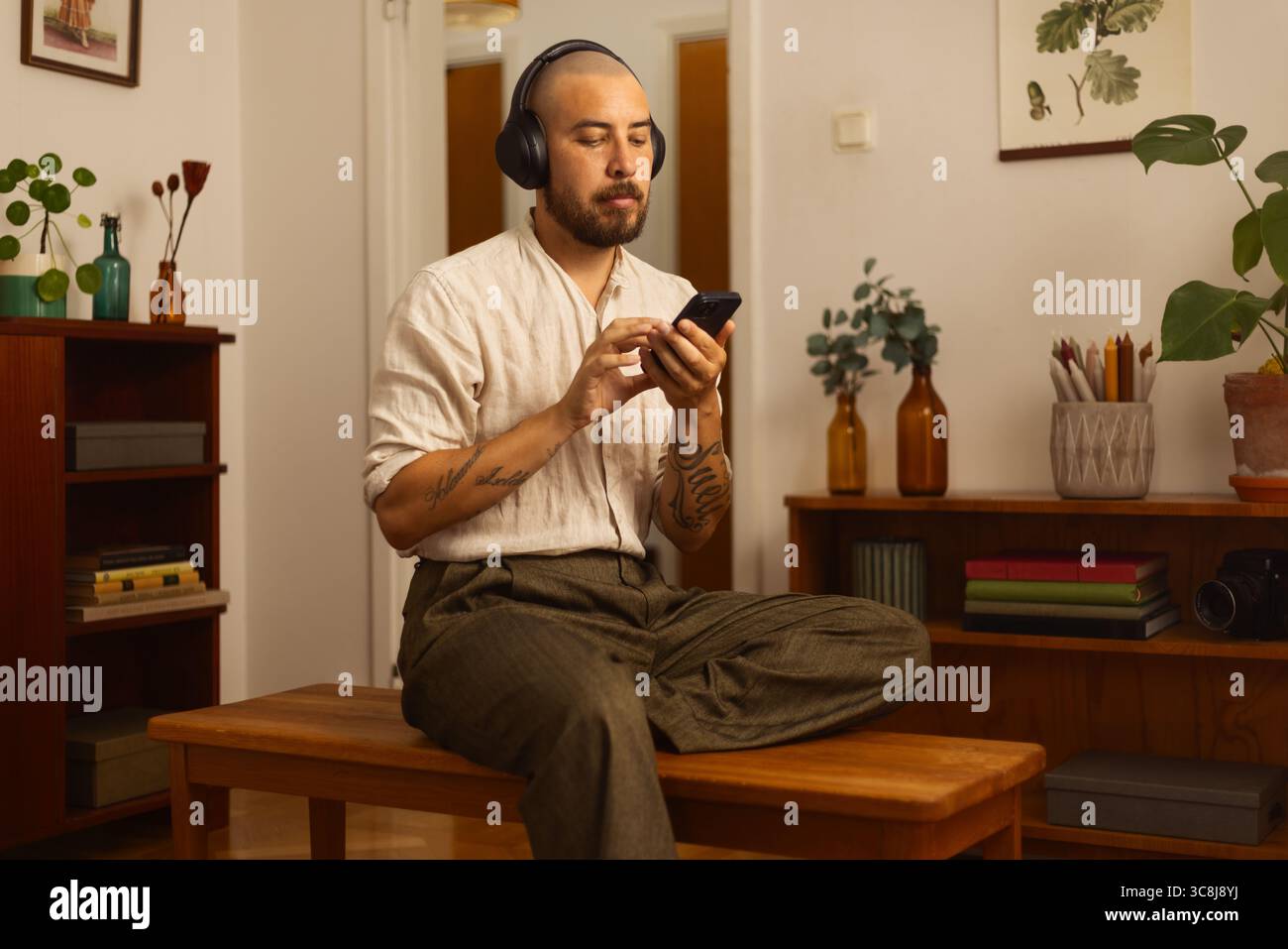 Uomo seduto a gambe incrociate su una panchina, con cuffie e smartphone in una stanza accogliente ed elegante. Un momento di calma di concentrazione digitale e relax. Foto Stock