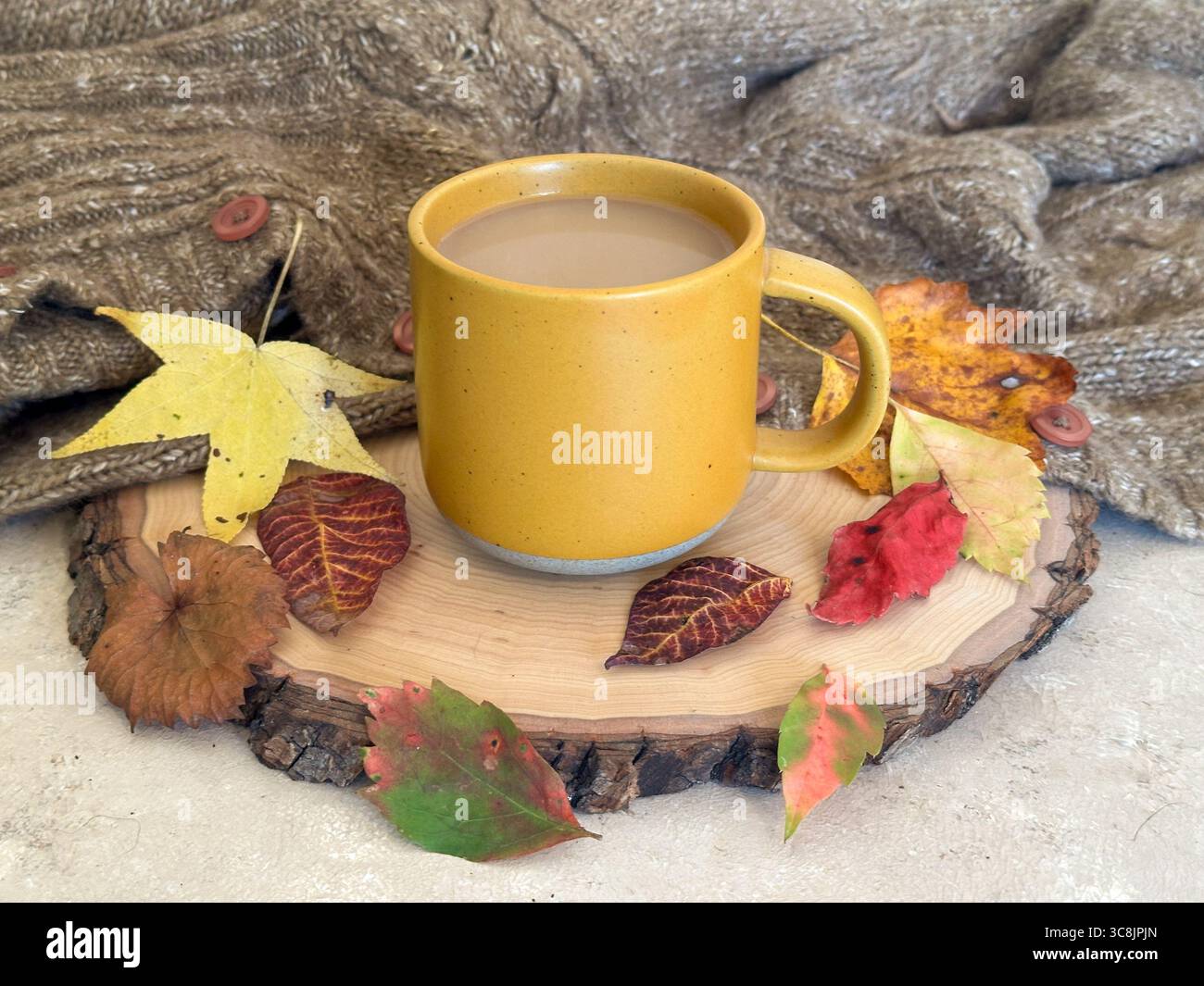 Una tazza di caffè gialla calda riempita di caffè si trova su una fetta di legno rustica, circondata da foglie autunnali colorate e da un comodo maglione a maglia Foto Stock