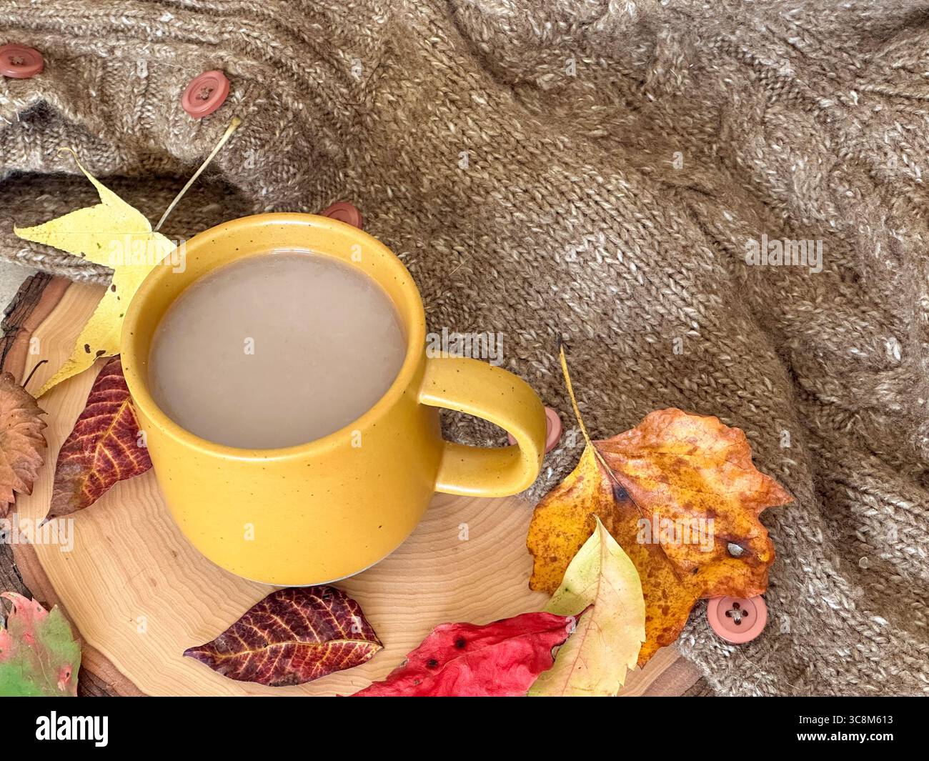 Un accogliente allestimento autunnale con una tazza di caffè gialla su una fetta di legno rustica, circondata da foglie autunnali colorate e un maglione a maglia Foto Stock