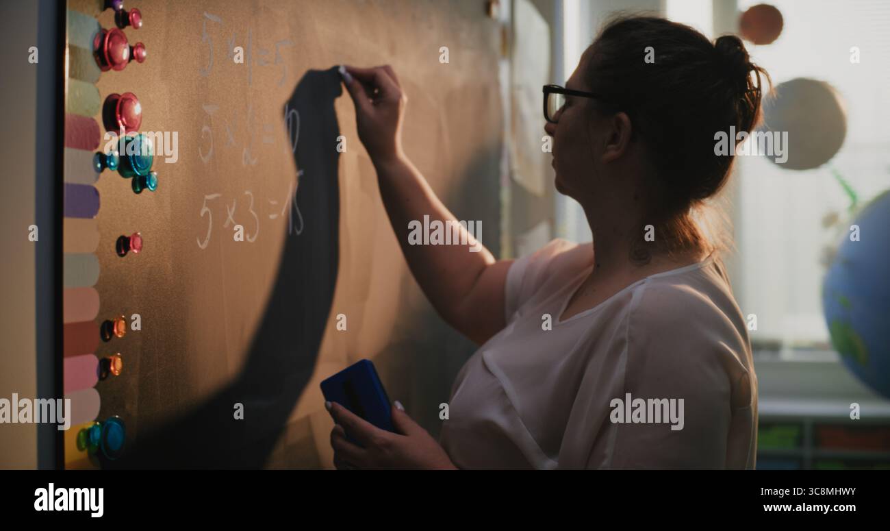 Insegnante donna che scrive numeri e tabella di moltiplicazione sulla lavagna, insegnare agli studenti delle scuole primarie a risolvere problemi di matematica durante la lezione. Corso di matematica in aula colorata. Primo piano. Foto Stock