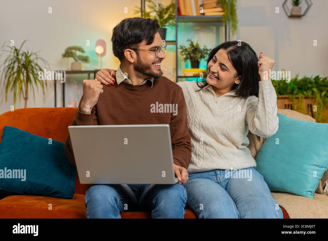 Allegro ha lasciato diverse coppie in abiti casual usando il computer portatile sorpreso con il jackpot di gioco online della lotteria che ha vinto un'e-mail sul divano nel soggiorno di casa. La famiglia indiana felice stringe i pugni in appartamento Foto Stock