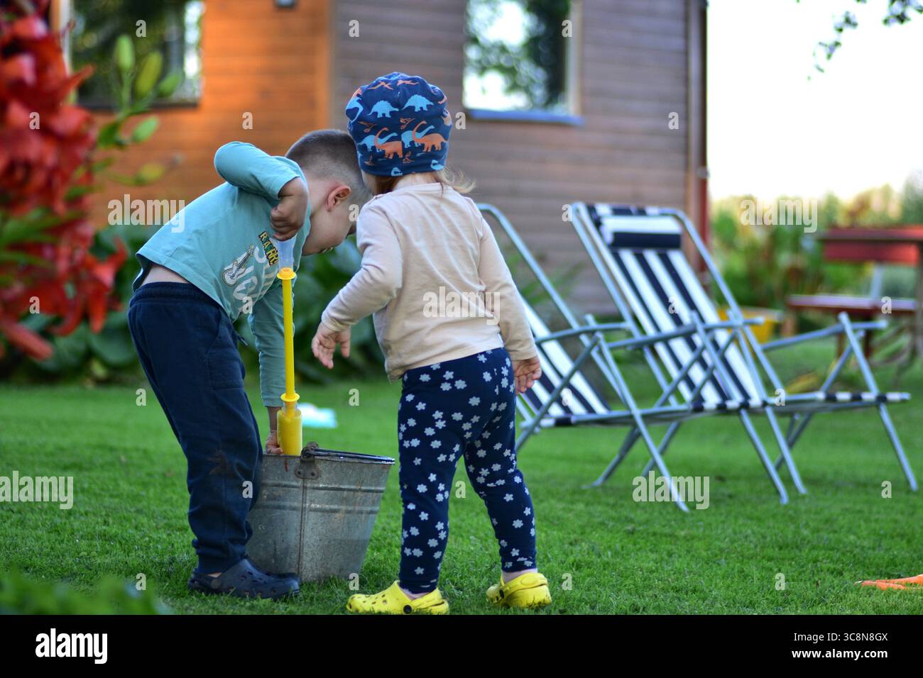 Due bambini che giocano con un secchio di metallo e un giocattolo ad acqua sul prato. I bimbi e i bambini esplorano la pompa dell'acqua insieme in una giornata estiva vicino a sedie e garde Foto Stock