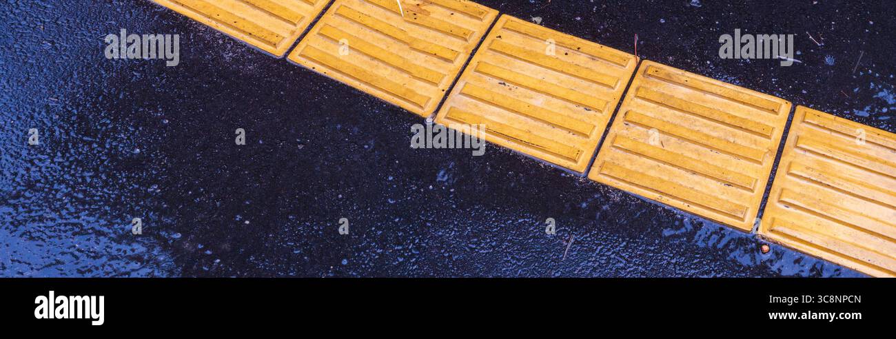 La pavimentazione tattile gialla in mattonelle di cemento con scanalature parallele su asfalto scuro bagnato aiuta i pedoni non vedenti e non vedenti a navigare Foto Stock