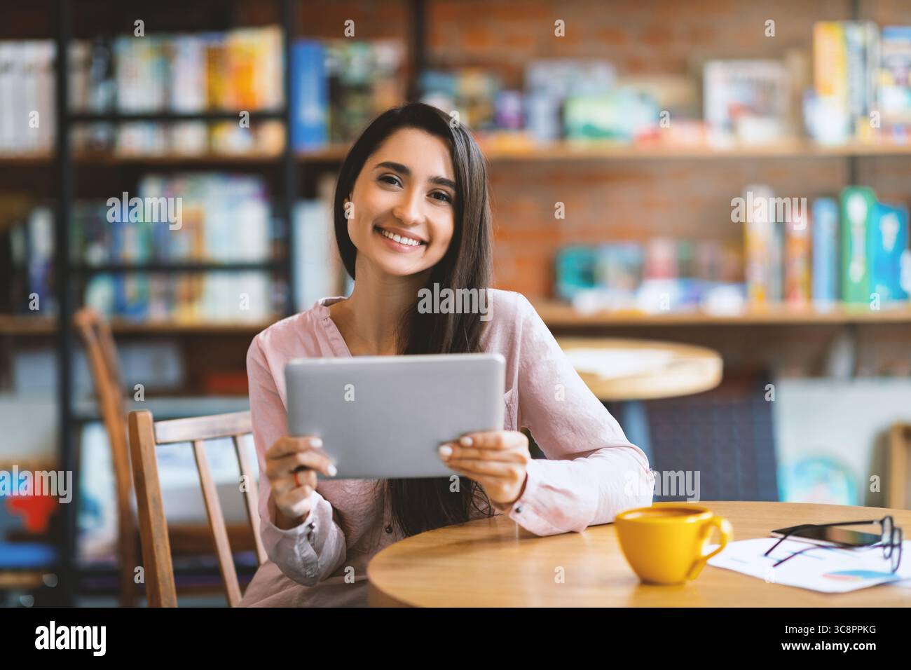 Donna araba felice seduta al bar, che usa un tablet digitale e beve caffè, sorride alla macchina fotografica, copia spazio Foto Stock