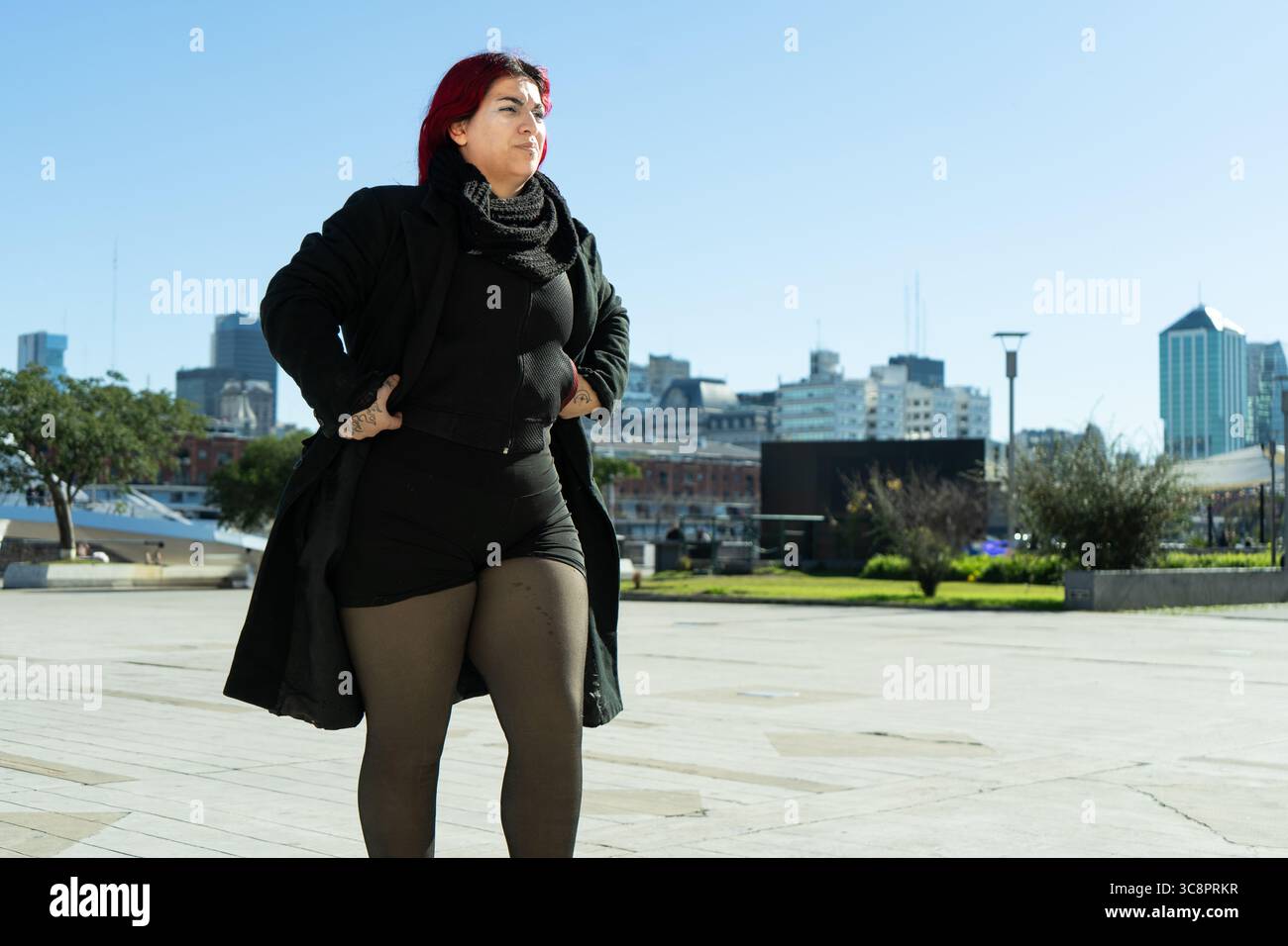 Elegante donna di grandi dimensioni con capelli rossi e tatuaggi posa con sicurezza con le mani sui fianchi in un ambiente moderno della città, mostrando la positività del corpo e Foto Stock