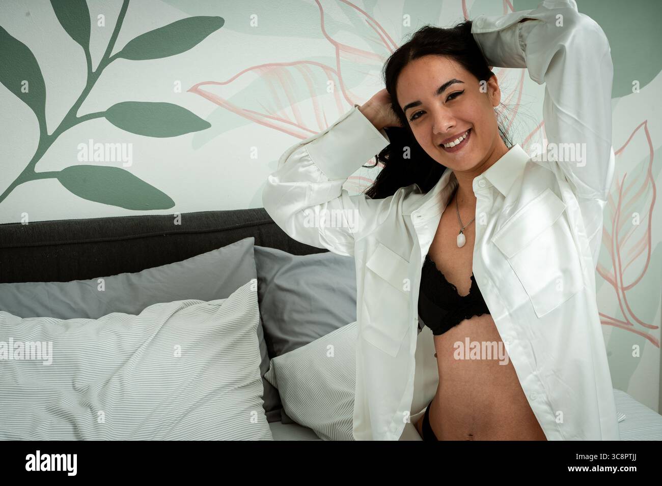 Donna sorridente che fissa i capelli mentre si siede sul letto in una camera da letto moderna con carta da parati floreale Foto Stock