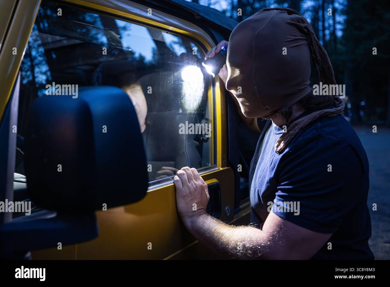 Un individuo mascherato emette una torcia elettrica attraverso il finestrino di un'auto mentre sbircia all'interno, suggerendo un tentativo di furto o di furto del veicolo. Foto Stock