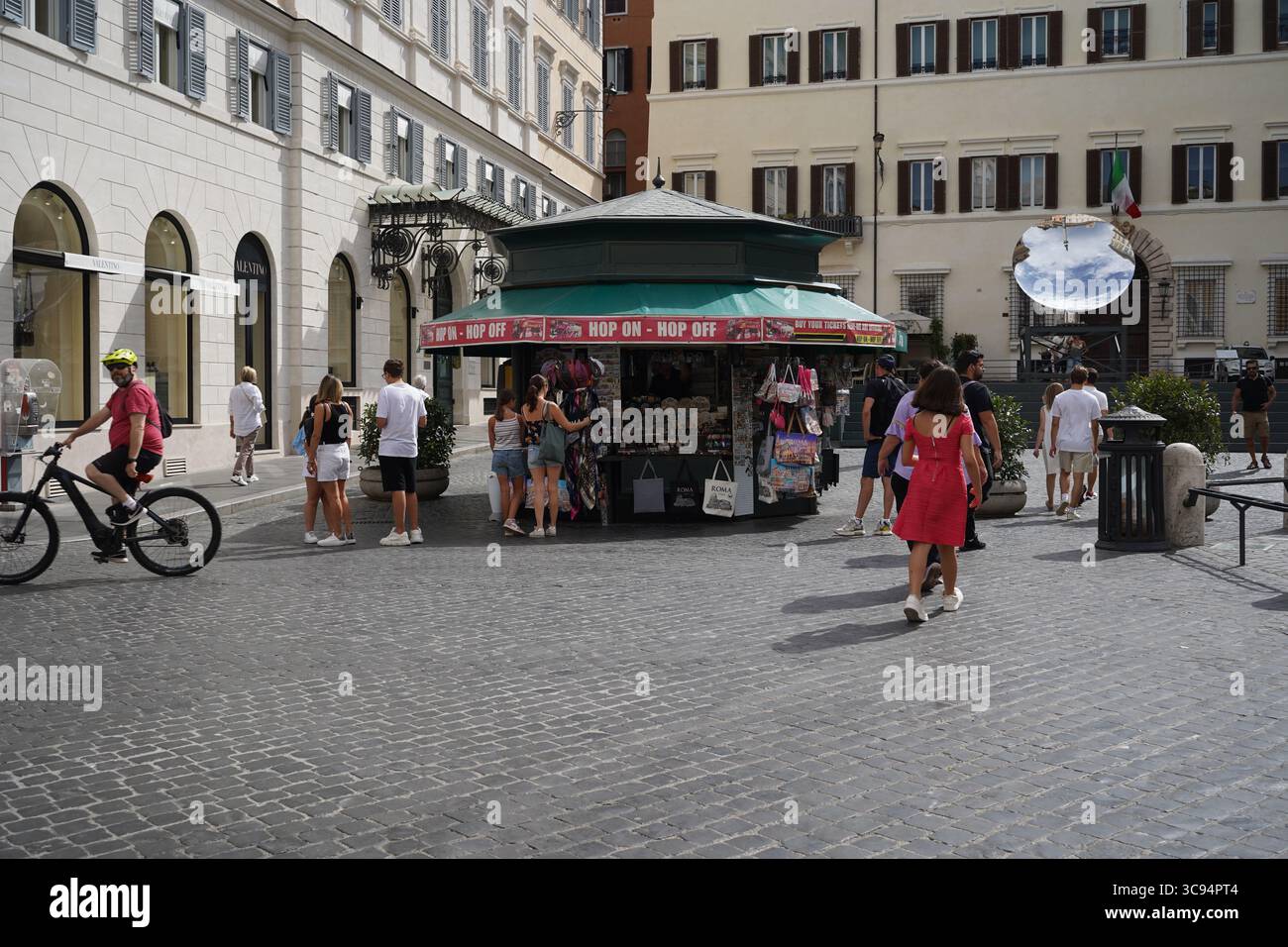 Persone intorno a un chiosco "Hop on Hop off" in Piazza di Spagna con un'installazione d'arte moderna e edifici storici sullo sfondo. Foto Stock