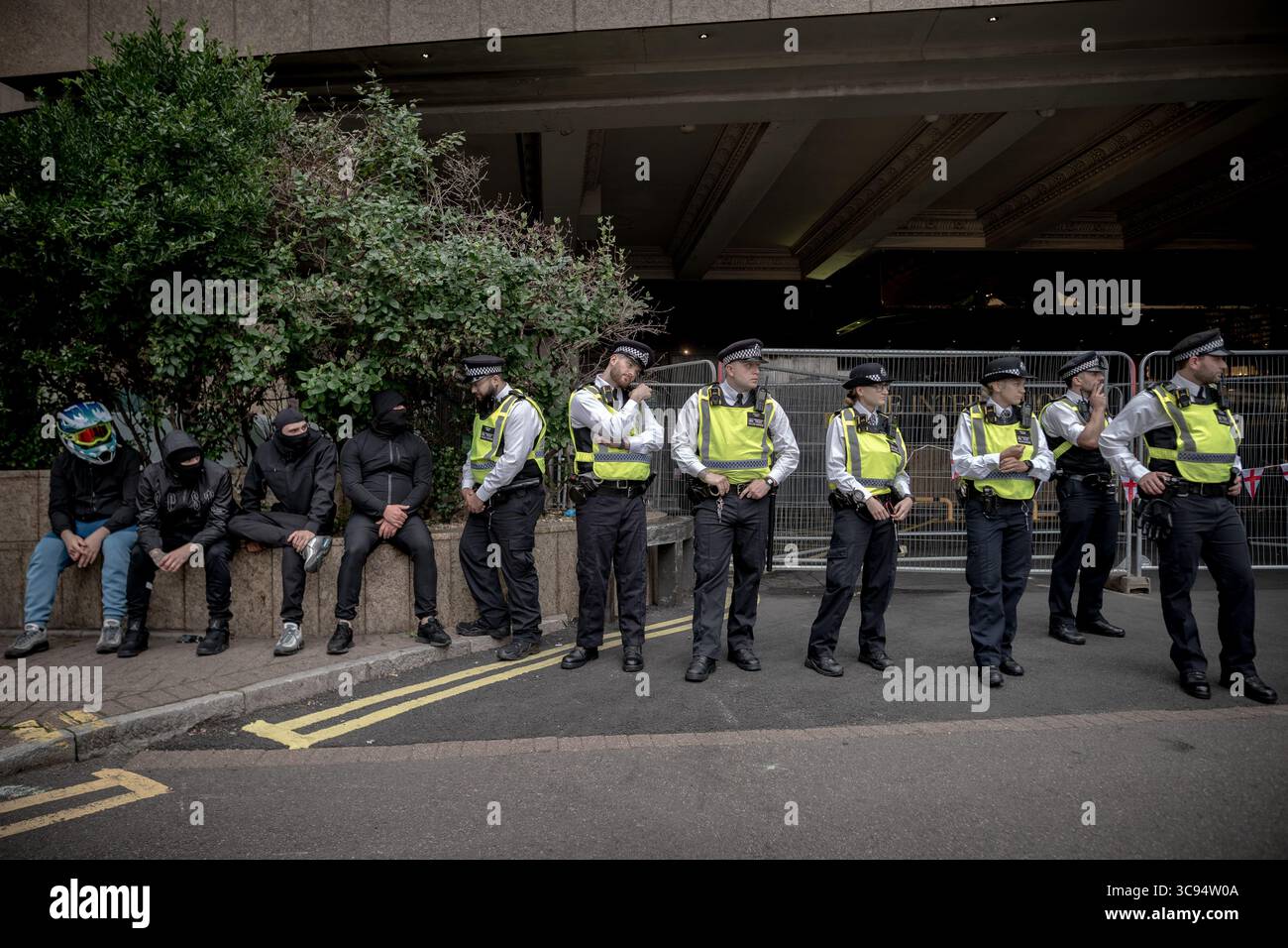 Londra, Regno Unito. 3 agosto 2025. Nella foto: La polizia Met sta di guardia fuori dall'hotel e guarda i manifestanti mascherati (L). I manifestanti anti-migranti manifestano al di fuori del Britannia International Hotel a Canary Wharf che attualmente ospita i richiedenti asilo. Un misto di uomini che indossavano maschere facciali con le famiglie che sventolavano bandiere e ascoltavano discorsi fuori dall'hotel a quattro stelle. Una serie di arresti sono stati fatti dalla polizia MET dopo che sono scoppiati scontri e scontri. Crediti: Guy Corbishley/Alamy Live News Foto Stock