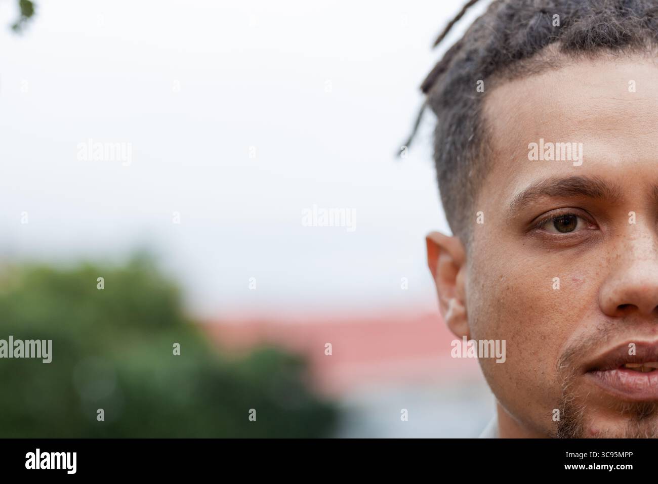 Uomo adulto che mostra mezza faccia con dreadlock in giardino accanto all'edificio con tetto rosso, spazio fotocopie Foto Stock