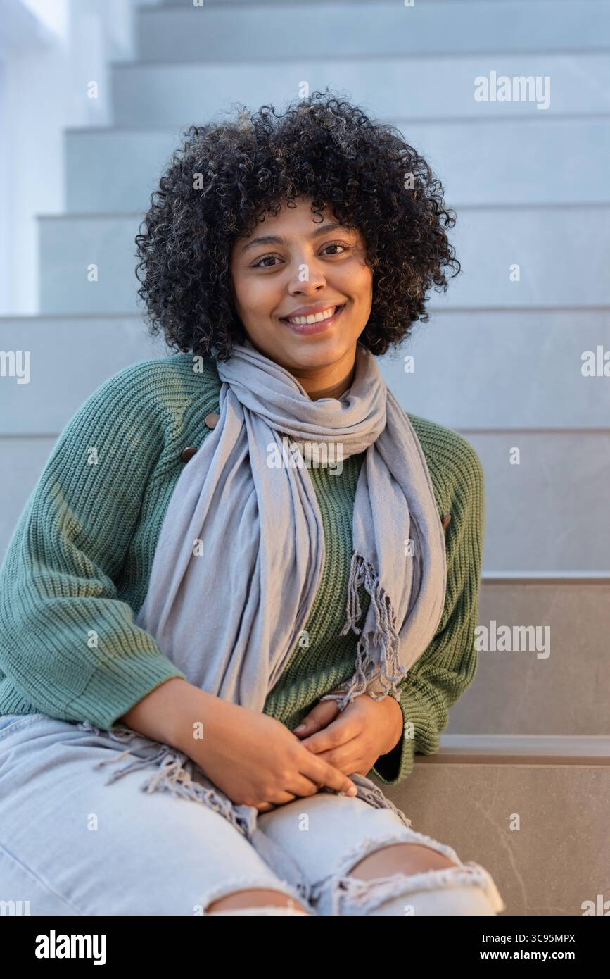 Donna afro-americana seduta su una scala con maglione verde, sciarpa grigia, jeans angosciati Foto Stock