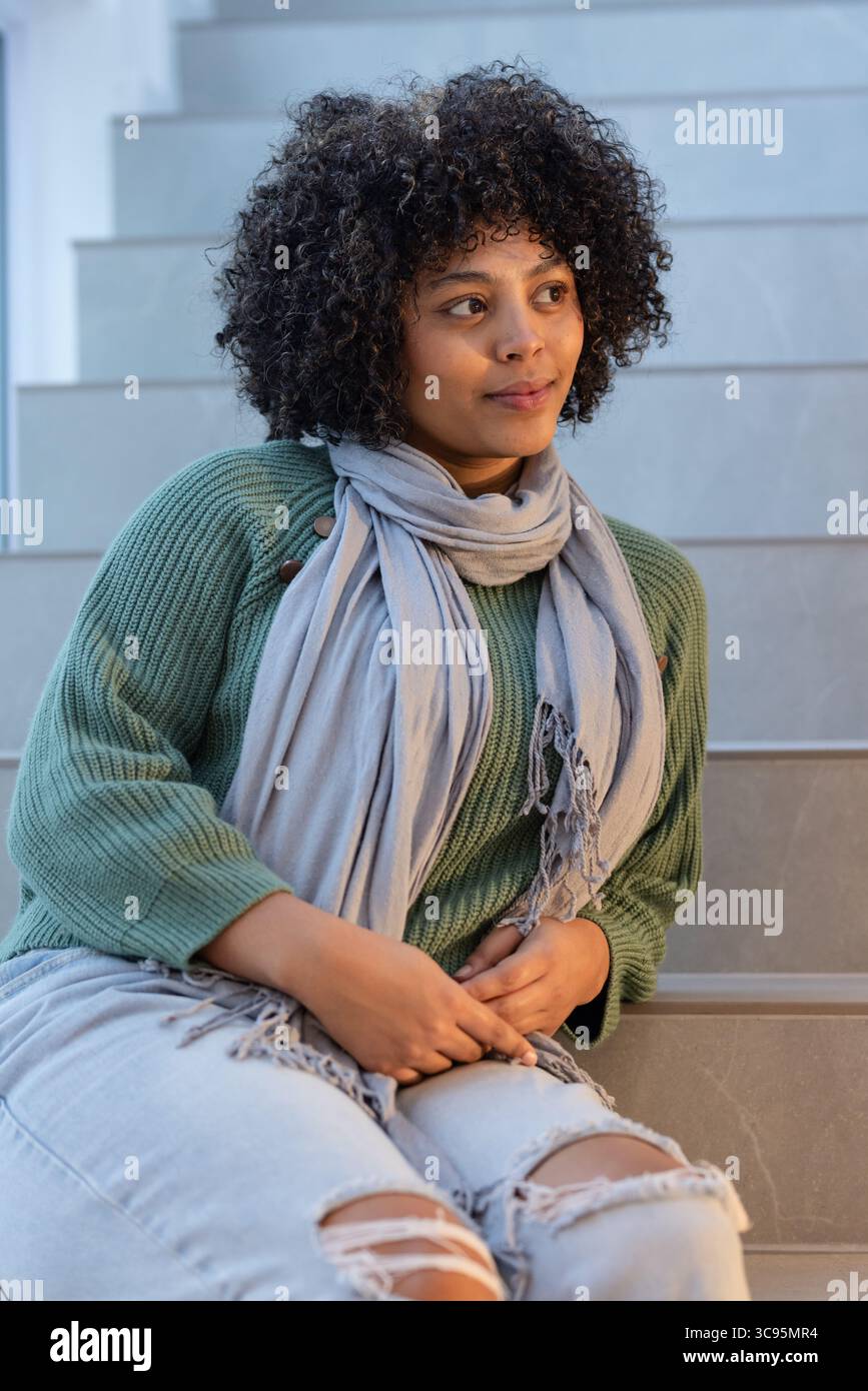 Donna afro-americana seduta su gradini grigi in scala con maglione verde e jeans strappati Foto Stock