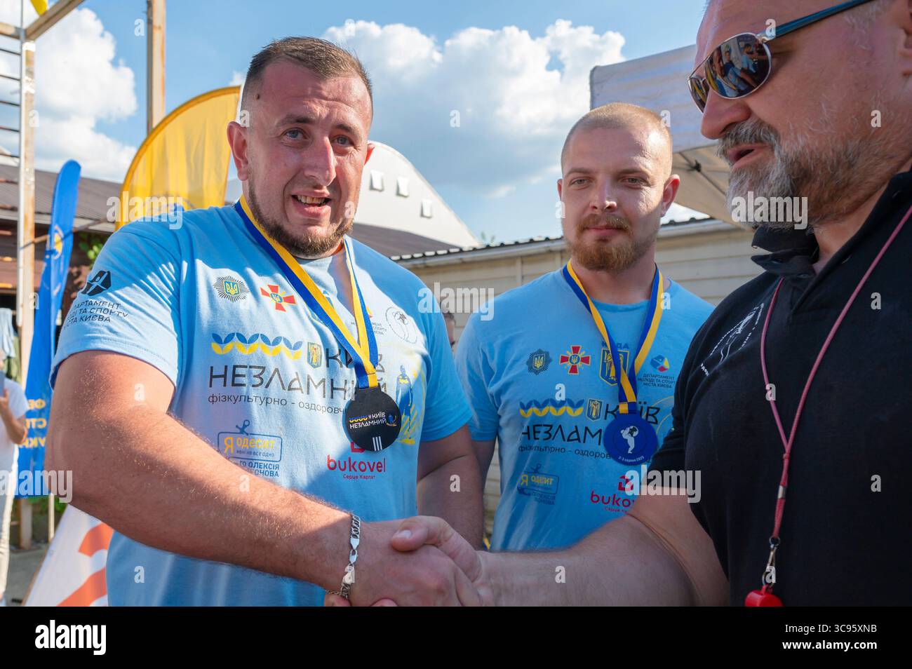 Kiev, Ucraina - 2d agosto, 2025:due veterani ucraini che indossano magliette blu con simboli e medaglie ucraini che stringono la mano dopo una competizione sportiva mentre un arbitro si congratula con loro Foto Stock