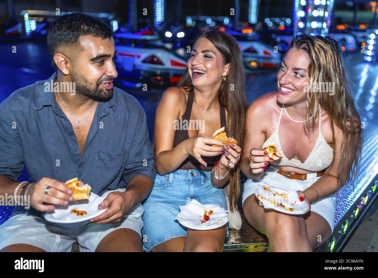 Tre amici si divertono con gli hot dog in un parco divertimenti, ridendo e divertendosi Foto Stock