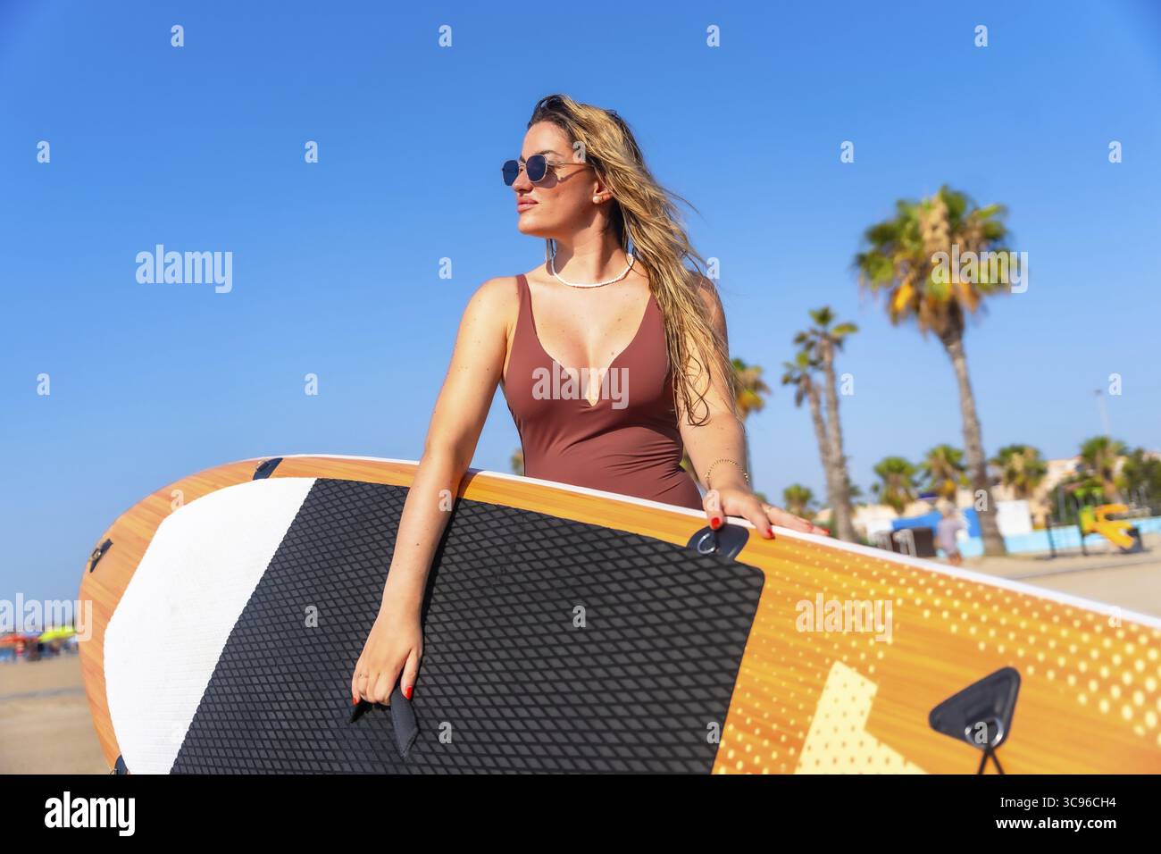 Surfista che porta la sua tavola da surf a pagaia lungo la spiaggia sabbiosa, godendosi una soleggiata giornata estiva piena di onde oceaniche e vibrazioni tropicali Foto Stock