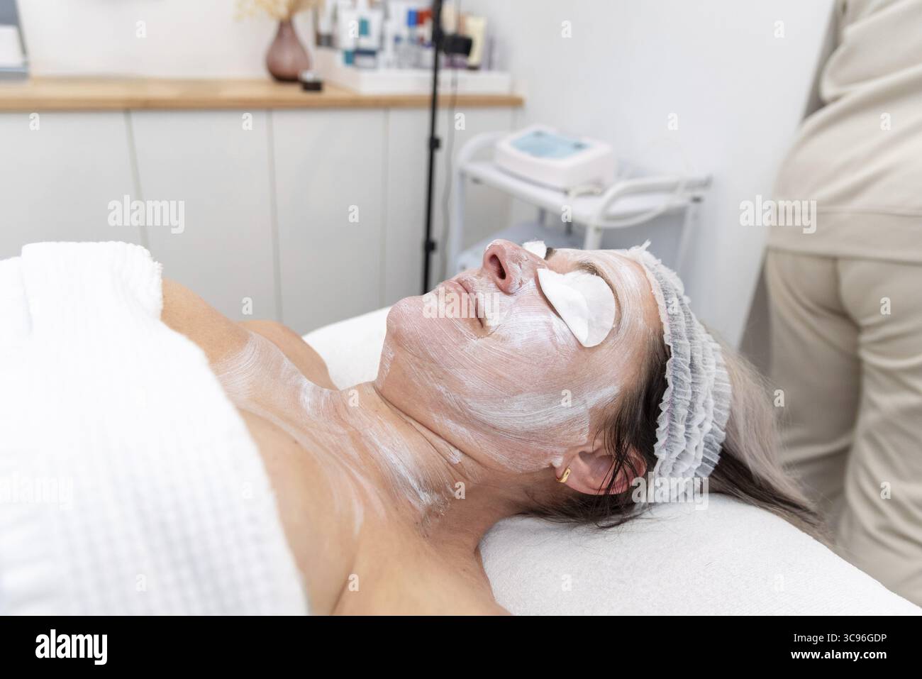 Donna matura che si gode il relax durante una procedura cosmetica facciale in un moderno salone di bellezza, circondato da un ambiente rilassante e cura professionale Foto Stock
