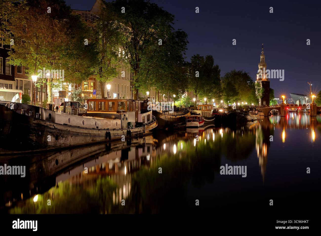 La torre di Montelbaanstoren e le verdi Oudeschans costeggiate dai canali di Amsterdam riflettono le luci incandescenti sull'acqua calma in un'affascinante scena fotografica notturna. Foto Stock