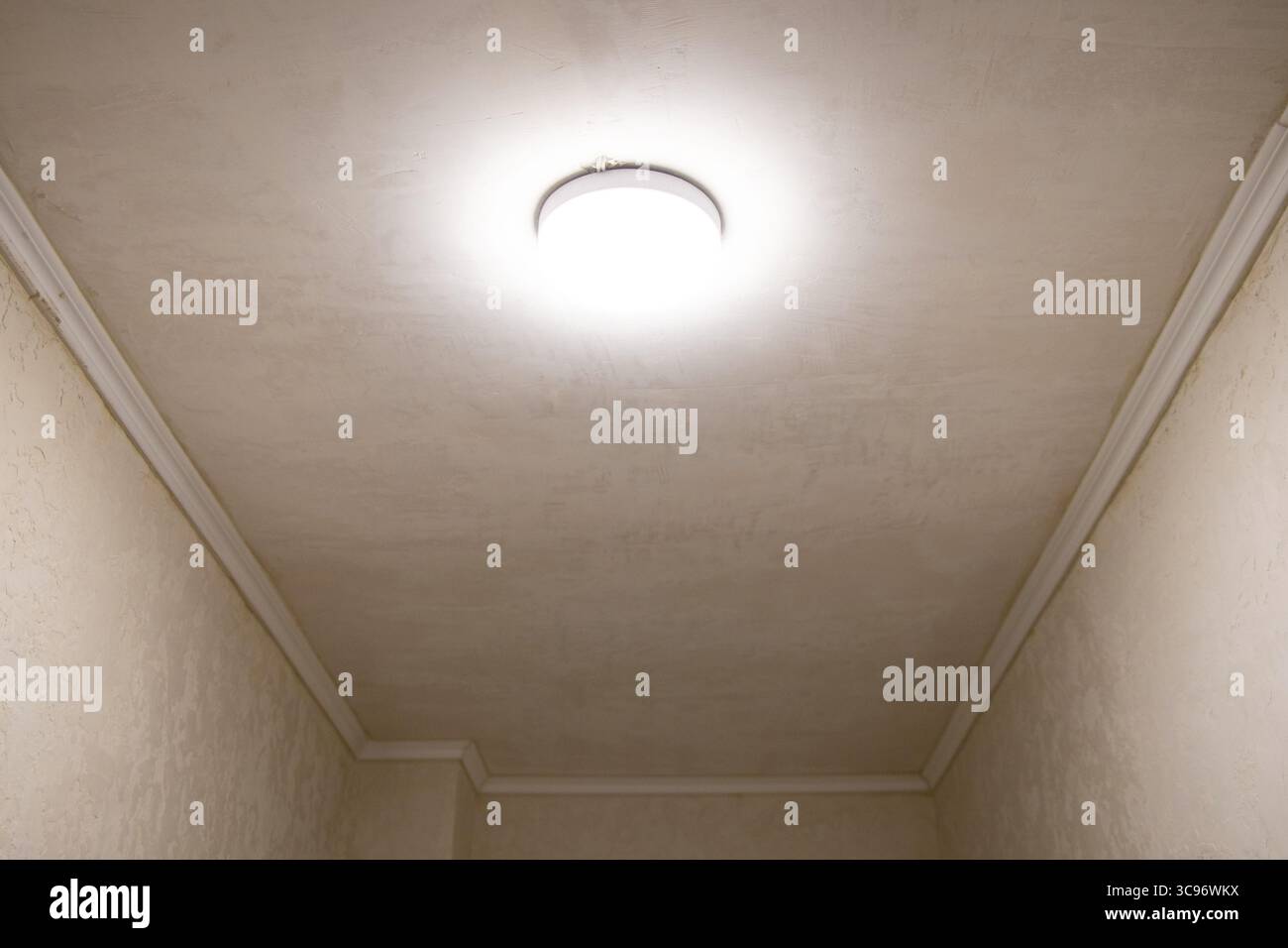 Lampadario luminoso sul soffitto di uno stretto corridoio con pareti beige e stucco, interni di appartamenti con illuminazione e finiture decorative Foto Stock