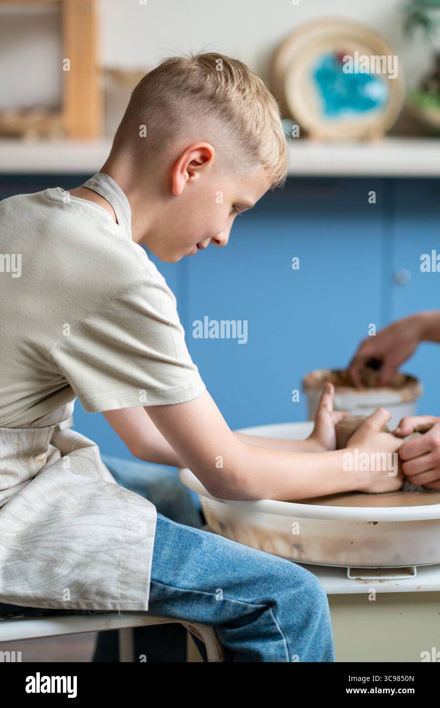 I ragazzi biondi imparano la ceramica, primo piano delle mani che modellano l'argilla su una ruota, attività creative per i bambini in un accogliente studio di ceramica, lavoro di squadra e abilità manuali Foto Stock