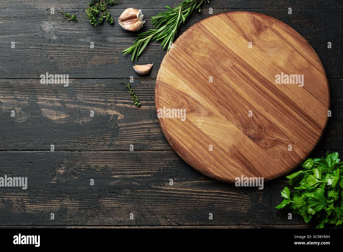 Lavagna rotonda in legno vuota con erbe e aglio su sfondo di legno scuro, vista dall'alto, spazio per copiare. Piatto di pizza in legno. Sfondo cibo, modello di progettazione Foto Stock