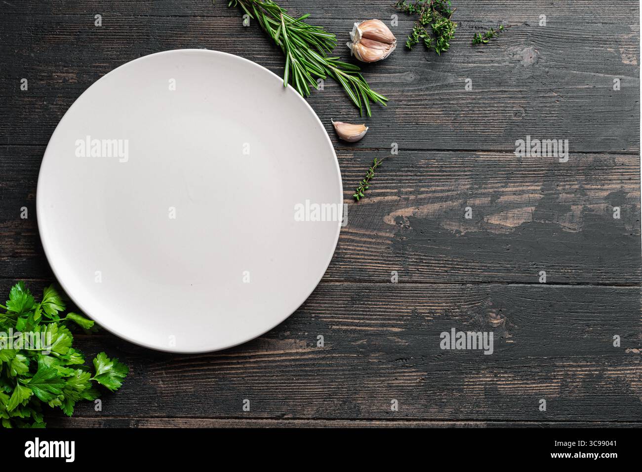 Piastra bianca vuota in ceramica con erbe e aglio su sfondo di legno scuro, vista dall'alto, spazio per copiare. Mockup da portata. Sfondo alimentare, design templ Foto Stock