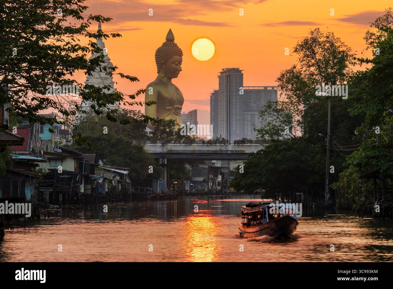 Una barca stava passando davanti alla statua del grande Buddha (Phra Buddha Dhammakaya Thepmongkhon) nel tempio Wat Pak Nam Phasi Charoen situato vicino al fiume durante il tramonto. Foto Stock