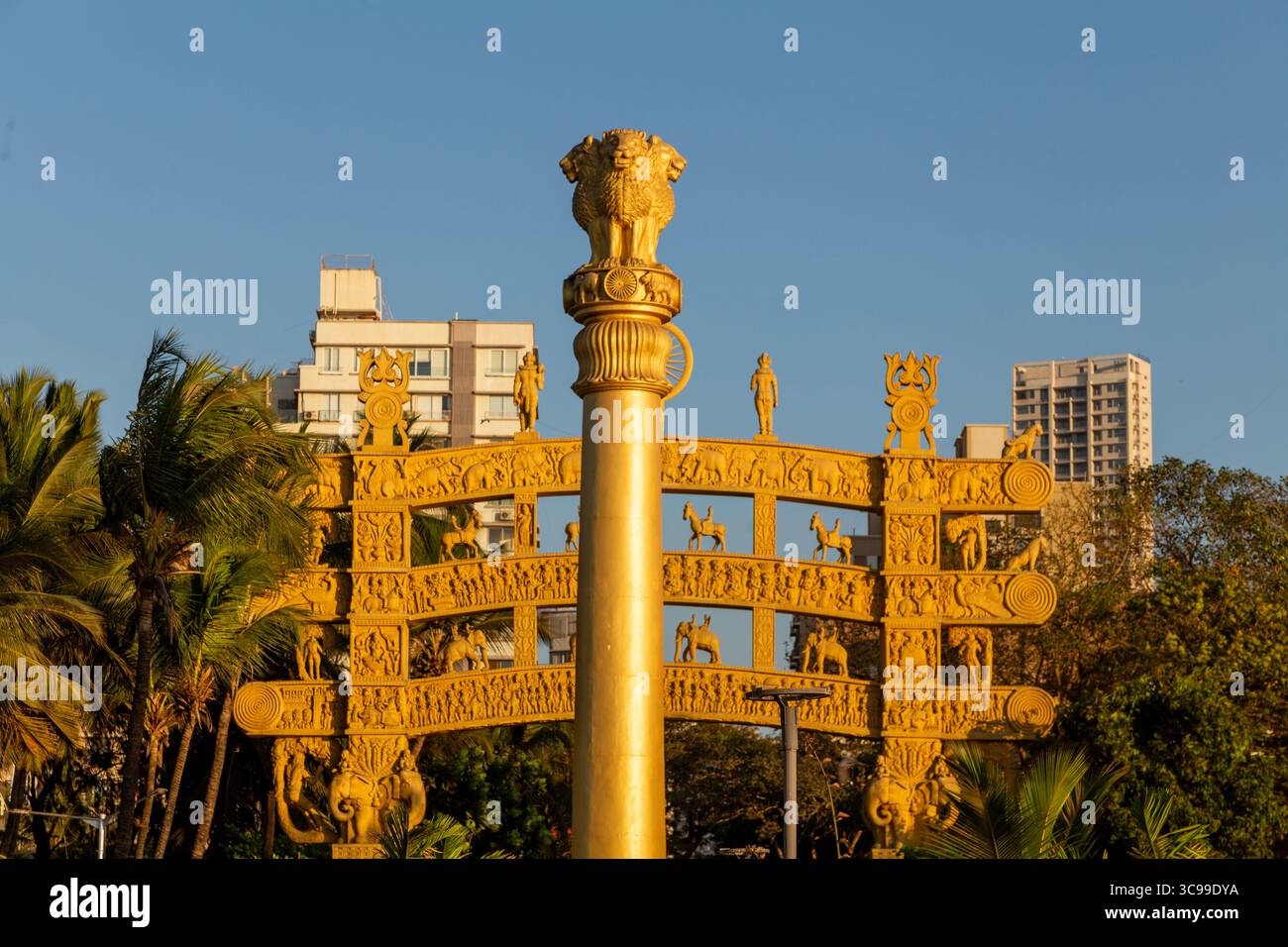 La colonna Ashoka d'oro e la porta ornata ispirata allo Stupa Sanchi si stagliano su un cielo azzurro, circondato da palme e dalla città moderna Foto Stock