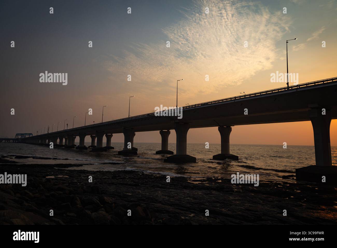 Un ponte moderno con alti pilastri si estende sul mare al tramonto, sagomato contro un cielo luminoso con nuvole spettacolari Foto Stock
