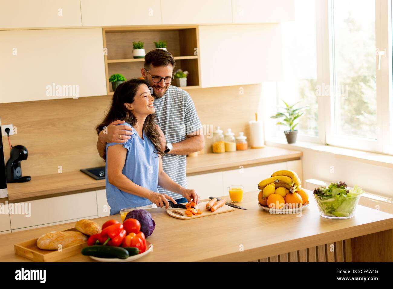 Una coppia condivide un momento caldo e divertente in una cucina soleggiata mentre trita ingredienti freschi, circondati da frutta e verdura vibranti. Il loro bon Foto Stock