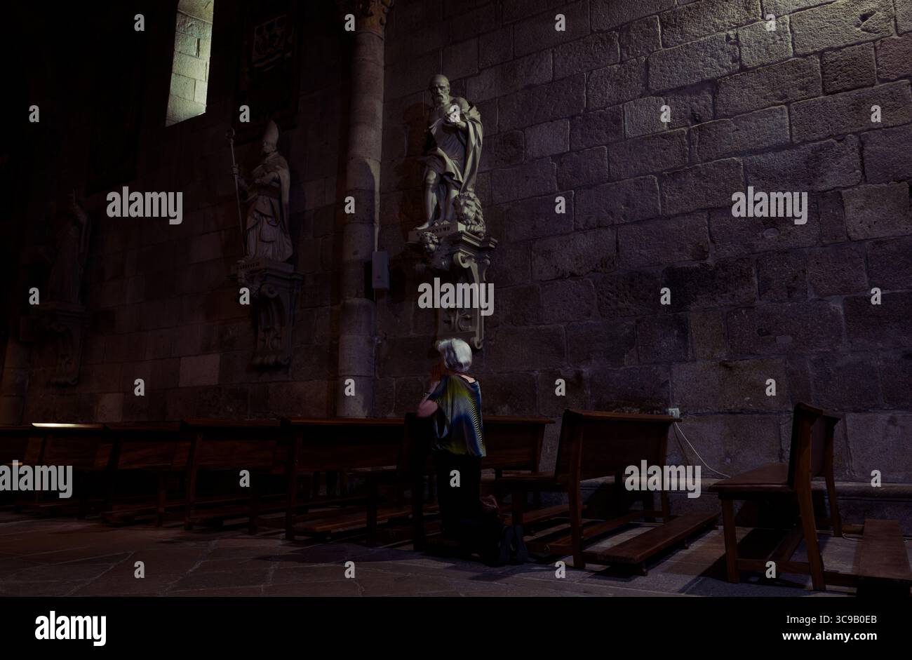 Donna inginocchiata in quieta preghiera sotto solenni santi in pietra in una chiesa poco illuminata a Braga Foto Stock