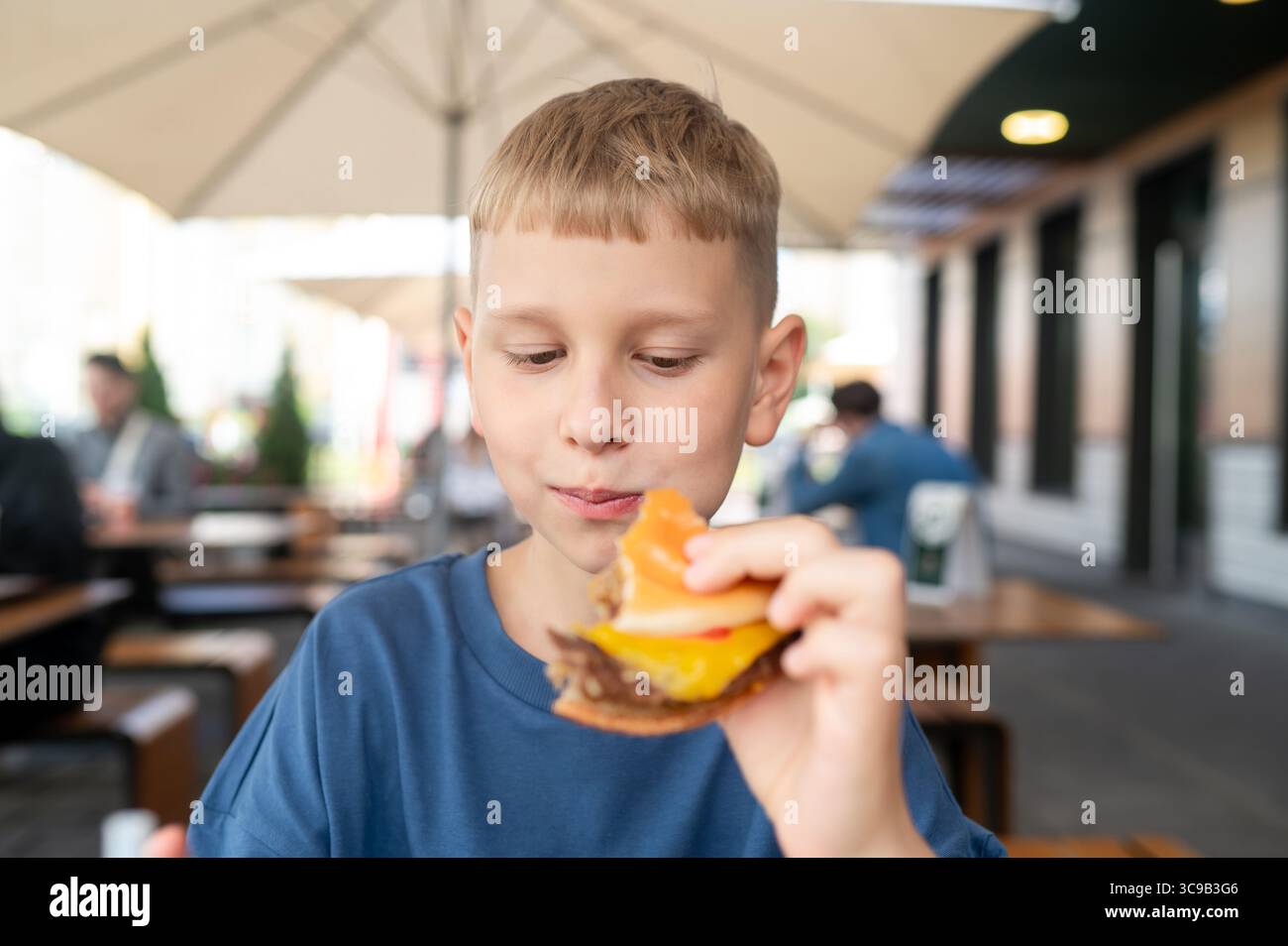 Momento informale di un ragazzino biondo seduto in una caffetteria all'aperto in una giornata di sole, tenendo un hamburger e godendo delle vibrazioni estive Foto Stock Momento informale di un ragazzino biondo seduto in una caffetteria all'aperto in una giornata di sole, tenendo un hamburger e godendo delle vibrazioni estive Foto Stock