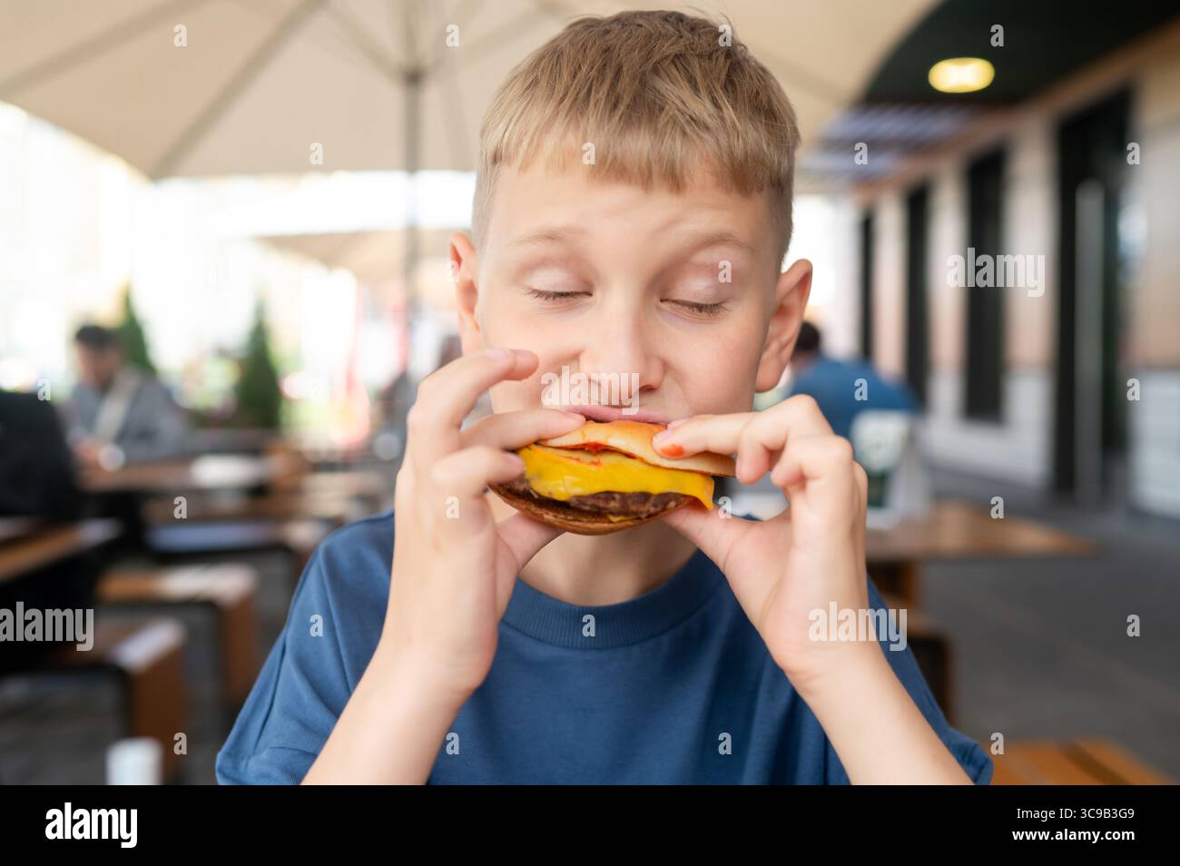 Momento informale di un ragazzino biondo seduto in una caffetteria all'aperto in una giornata di sole, tenendo un hamburger e godendo delle vibrazioni estive Foto Stock Momento informale di un ragazzino biondo seduto in una caffetteria all'aperto in una giornata di sole, tenendo un hamburger e godendo delle vibrazioni estive Foto Stock