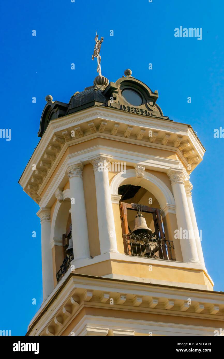 Una vista dettagliata di un campanile storico della chiesa con colonne bianche, un'iscrizione del 1881 e una croce dorata contro un cielo blu brillante. Foto Stock