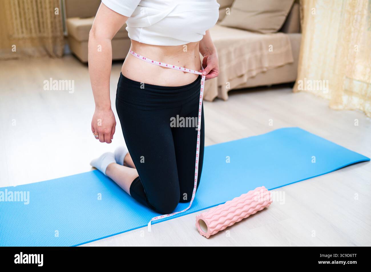Donna con vita imperfetta che lo misura con il nastro mentre sta in piedi su un tappetino yoga, consapevolezza del corpo e concetto di cura personale Foto Stock