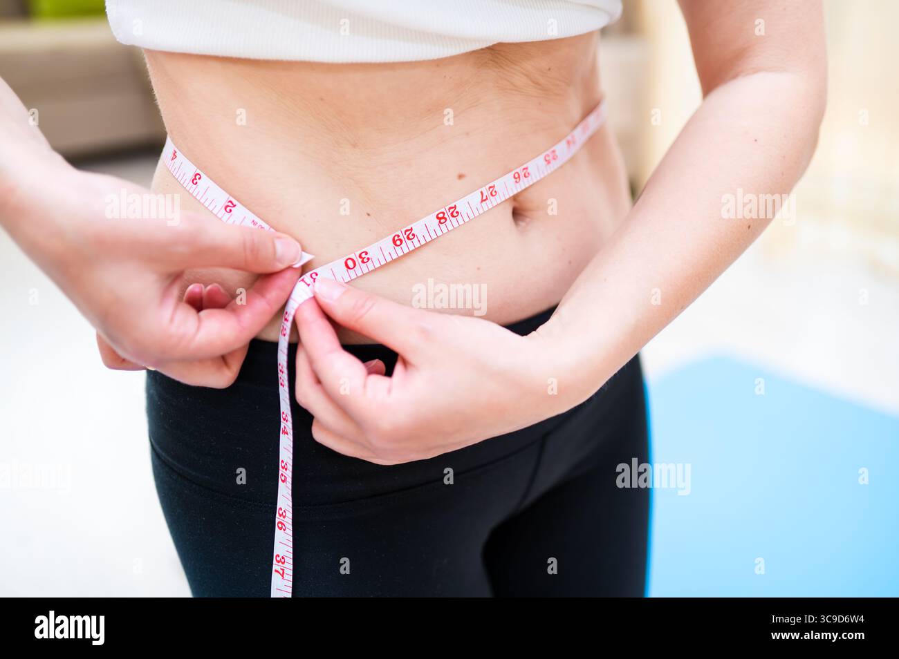 Donna con vita imperfetta che lo misura con il nastro mentre sta in piedi su un tappetino yoga, consapevolezza del corpo e concetto di cura personale Foto Stock