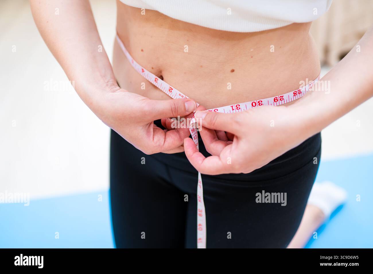 Donna con vita imperfetta che lo misura con il nastro mentre sta in piedi su un tappetino yoga, consapevolezza del corpo e concetto di cura personale Foto Stock