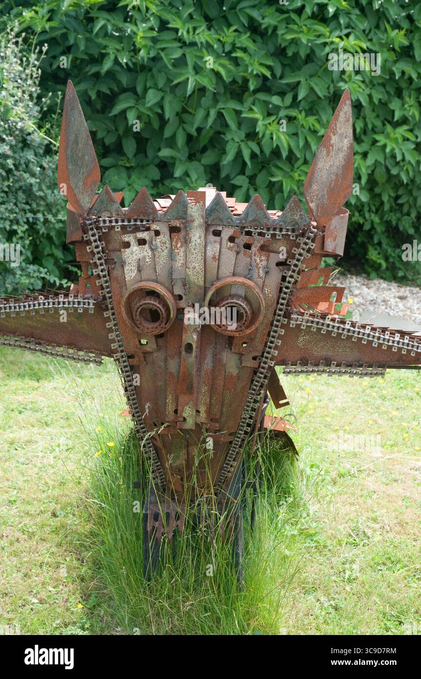 Un'impressionante scultura simile a un drago si erge in un giardino, realizzata in metallo riciclato e adagiata su un lussureggiante fogliame verde. Foto Stock