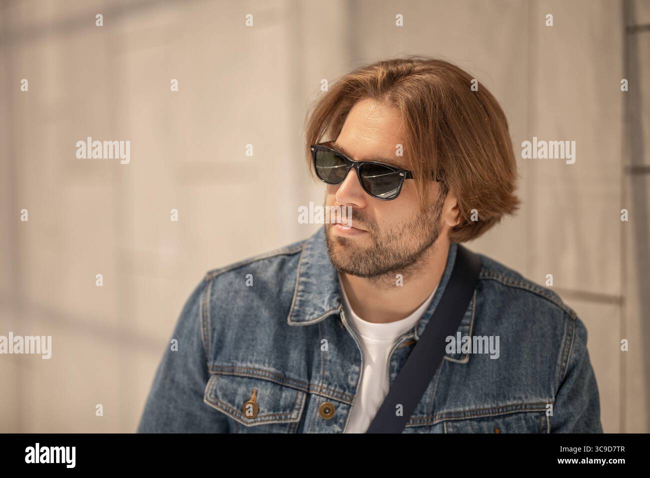 Un giovane elegante dai capelli lunghi indossa occhiali da sole, una giacca in denim e una T-shirt bianca in un ritratto casual. Perfetto per la moda, lo stile di vita e te Foto Stock