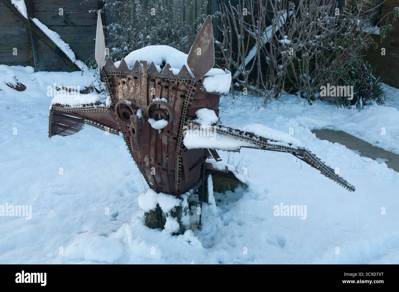 Scultura artistica in metallo a forma di creatura mitica si erge in un giardino innevato, circondato da piante invernali e luce mattutina. Foto Stock