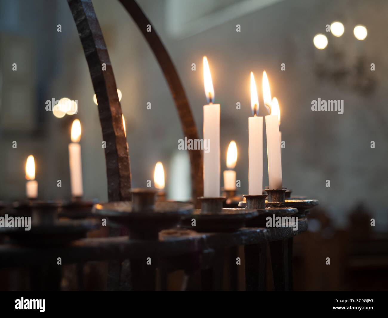 Candele accese su un supporto in ferro battuto all'interno di una chiesa, simboleggiano la preghiera, il ricordo e la pace, con luce soffusa e interni sacri sfocati Foto Stock