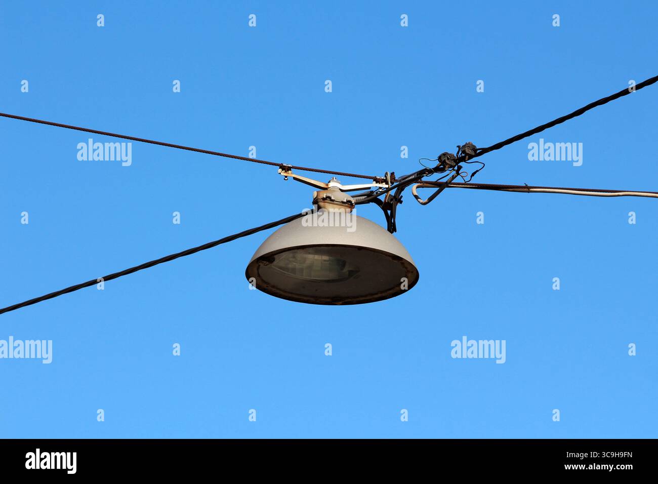 Una classica lampada da strada industriale è appesa ai cavi sospesi, al suo paralume metallico e all'hardware esposto visibile contro un cielo blu limpido, evocando la città Foto Stock