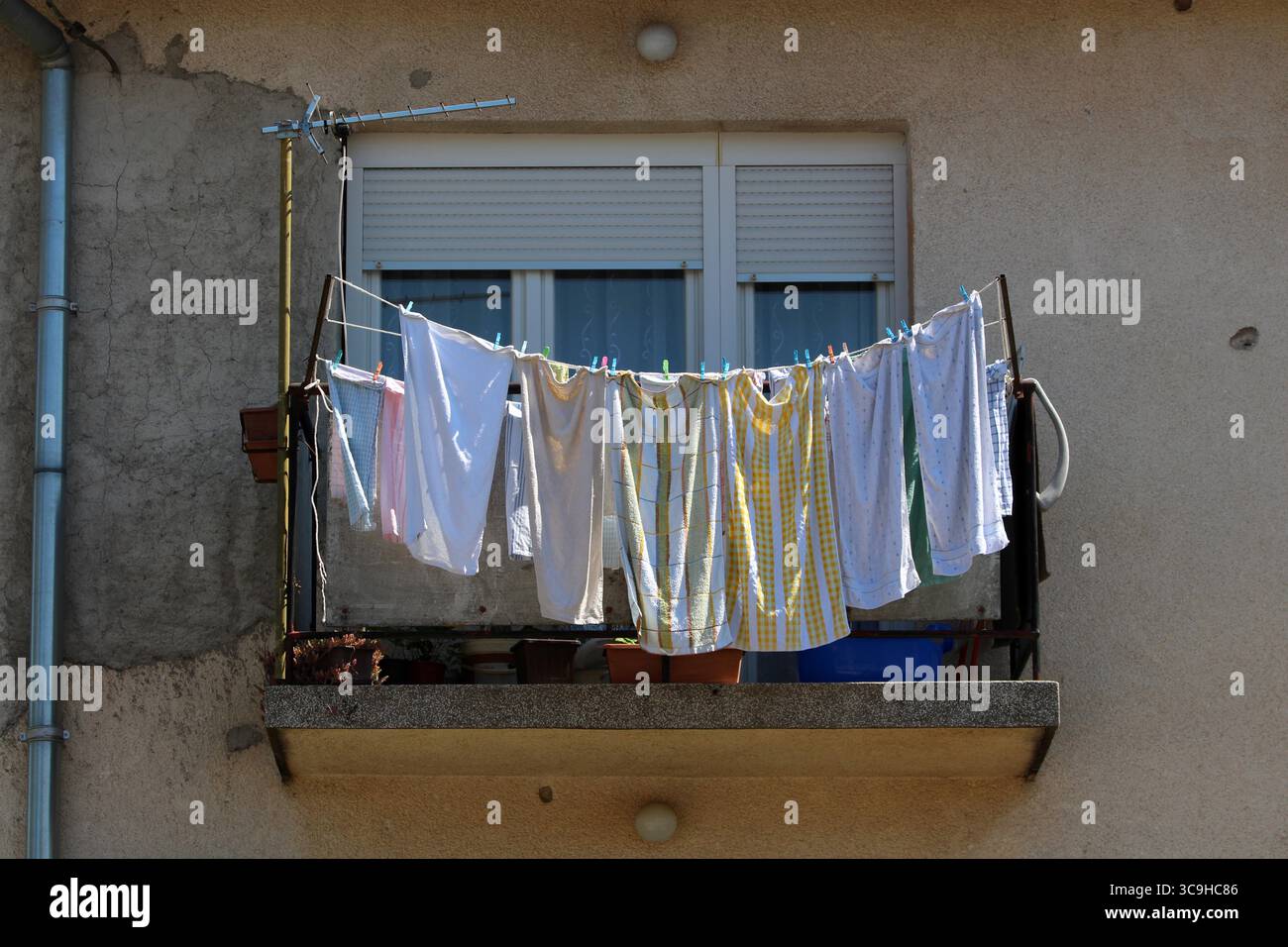 La lavanderia in vari colori e fantasie è appesa a una ringhiera del balcone, gettando ombre su cemento intempestivo e piante in vaso sotto una finestra Foto Stock