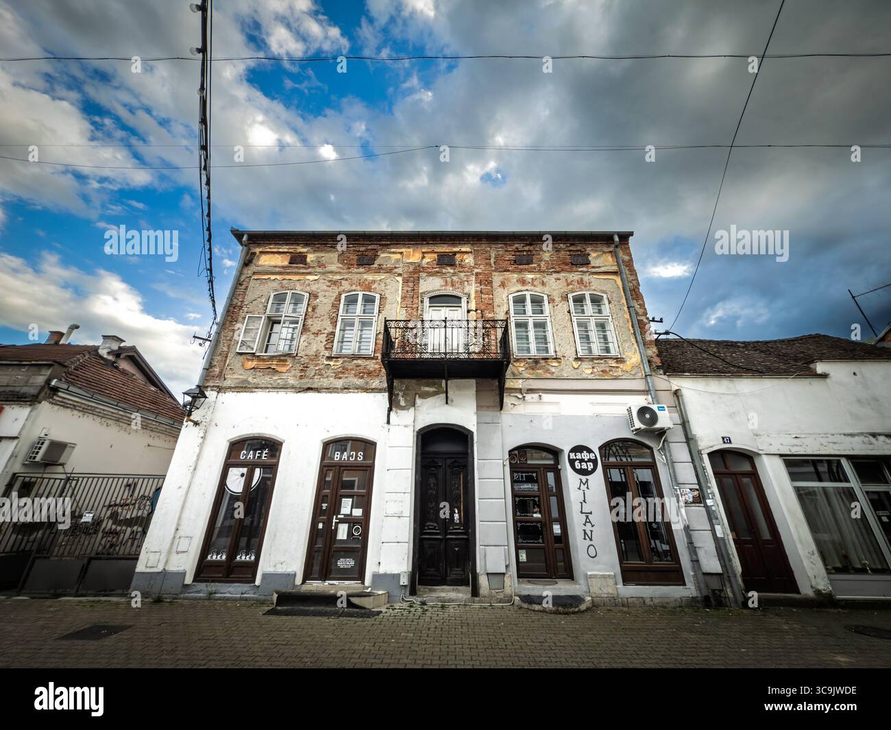 VALJEVO, SERBIA - 12 MAGGIO 2025: Una vecchia casa cittadina nel centro della città ospita piccoli bar a livello della strada. Mattoni esposti, intonaco pelato e una balla piegata Foto Stock