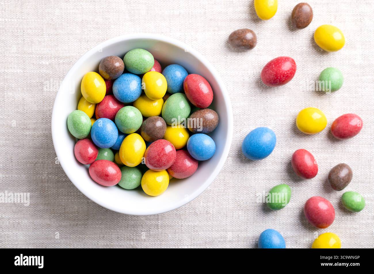Arachidi al cioccolato al latte ricoperte di caramelle, in una ciotola bianca, su tessuto di lino. Arachidi tostate, ricoperte di cioccolato al latte, ricoperte di zucchero multicolore. Foto Stock