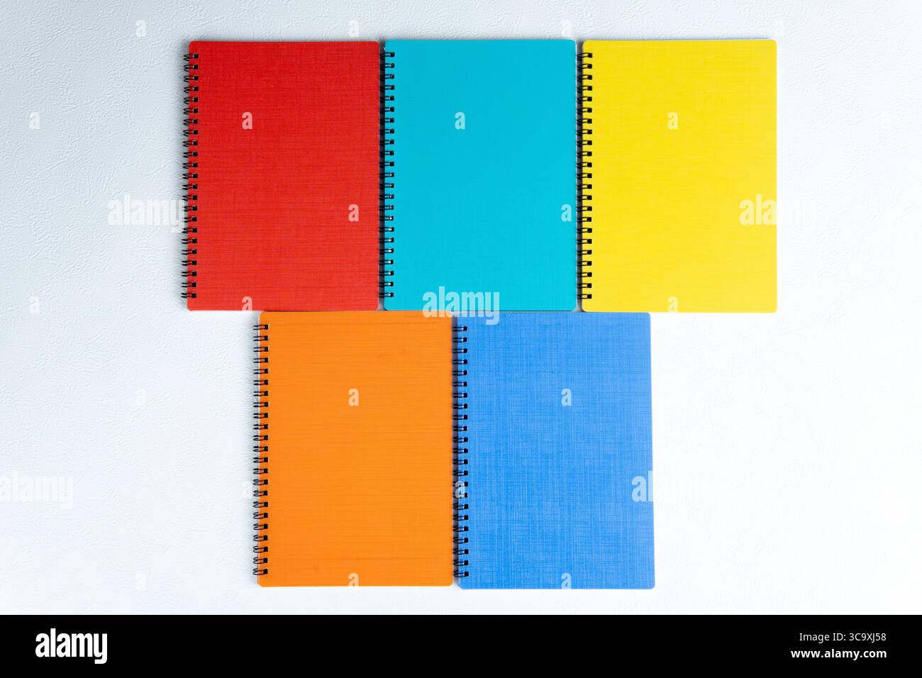 Vista dall'alto di cinque notebook a spirale in rosso, arancione, blu, giallo e verde acqua. Disposte su una superficie ruvida bianca. Foto Stock
