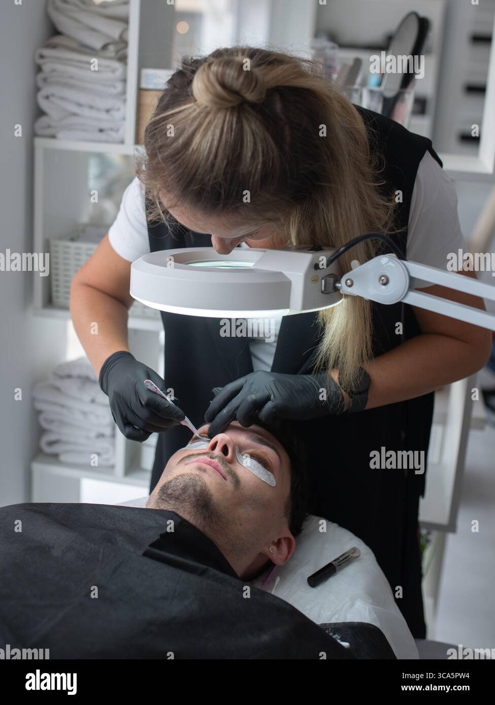 Il tecnico di bellezza sta applicando con cura le prolunghe per ciglia ai clienti maschi in un moderno salone, caratterizzato da un'illuminazione luminosa e da un ambiente di lavoro pulito e organizzato Foto Stock